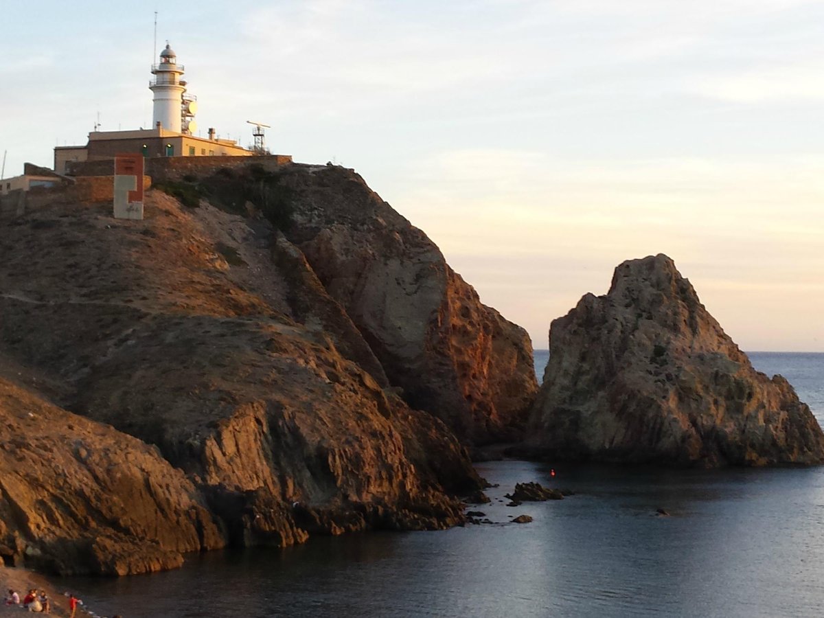 The iconic Faro de Cabo de Gata lighthouse on a rugged cliff overlooking the Mediterranean Sea in Almeria, with waves crashing against the rocky coastline.