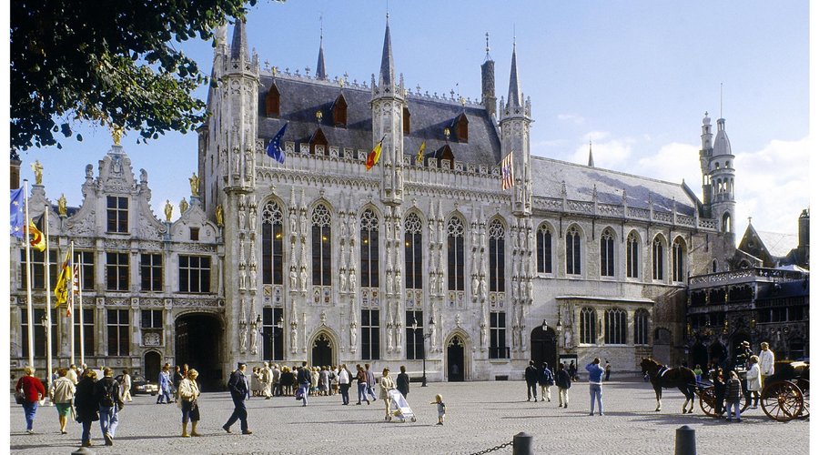 The ornate Gothic facade of the Bruges City Hall (Stadhuis) dominating the historic Burg Square, with tourists admiring the architecture.