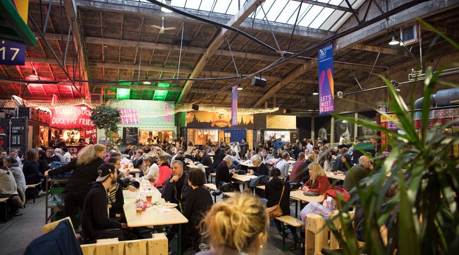 Aarhus, Denmark: Bustling interior of Aarhus Street Food market with diverse food stalls, glowing signs, and people enjoying meals at communal tables.