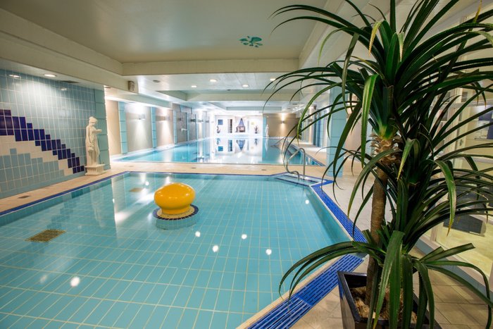 Indoor swimming pool with blue tiles and a statue, surrounded by plants.