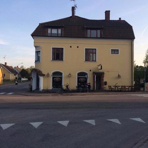 Pizzeria på bottenvåningen. Trafikerad väg på vänster sida och parkering på höger sida.