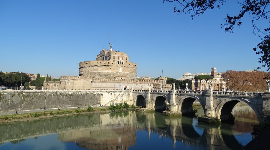 Castel Sant'Angelo in Rome, Italy, seen from across the Tiber River with Ponte Sant'Angelo leading to it, under a warm evening sky.