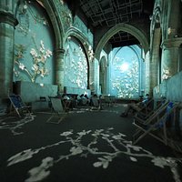 Projections cover the walls and the floor, dancing over visitors seated in socially-distanced deck chairs.