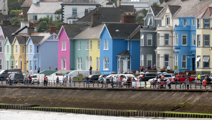 The charming promenade of Whitehead, Northern Ireland, stretching along the calm waters of Belfast Lough with boats moored and distant green hills under a clear sky.