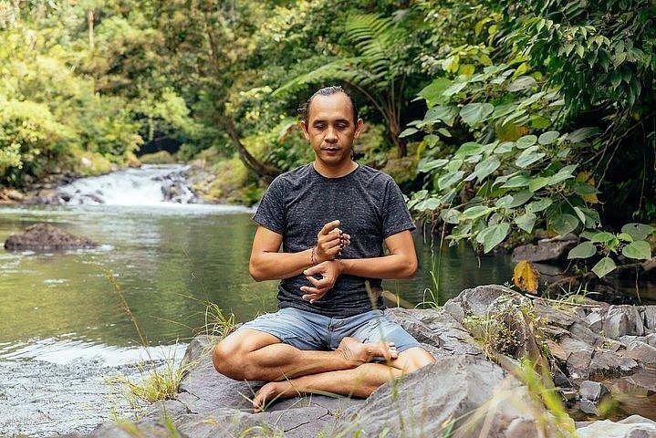 A person with eyes closed sitting in Lotus pose outside by a river.