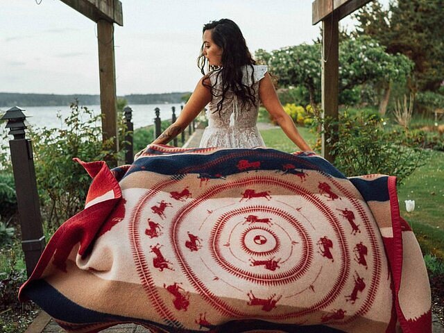 Maka Monture Päki in a white dress unfurling a Native American textile behind her.