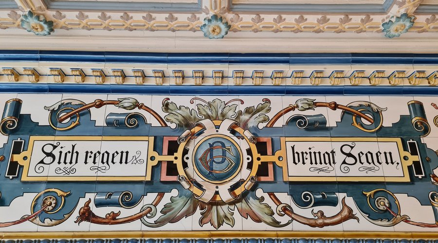 Dieses Bild zeigt einige der handbemalten Fliesen von Villeroy und Boch. In der Mitte des Spruchs befinden sich die Initialen GP für die Gebrüder Pfund.

This picture shows some of the hand painted tiles from Villeroy and Boch. In the middle of the saying are the initials GP for Gebrüder Pfund. 