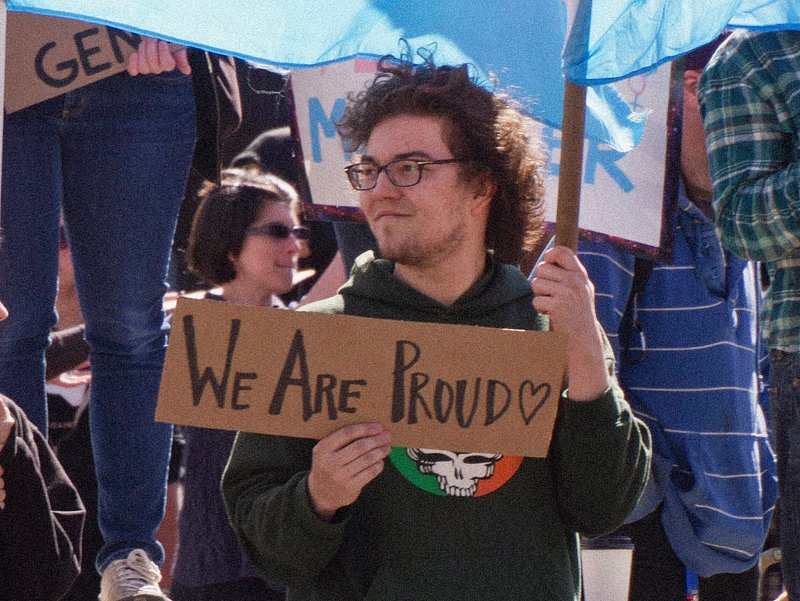 Person standing with 'We Are Proud' sign and holding a Transgender Pride flag