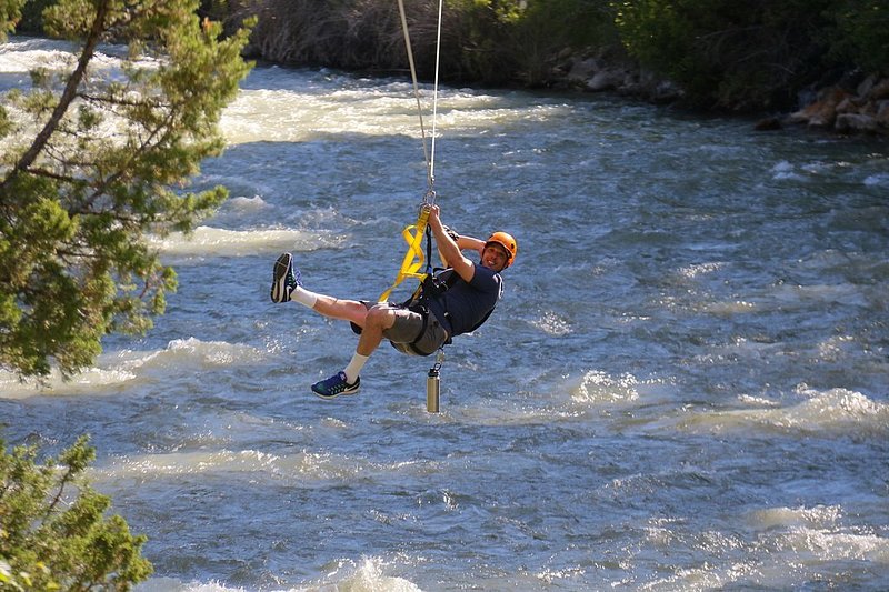 A smiling man ziplines across the gurgling Gallatin River.