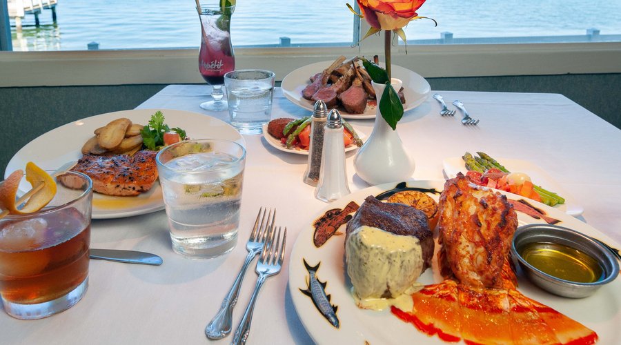 A full table with a waterfront view, boasting a hearty plate of Surf & Turf, Grilled Salmon with Fingerling Potatoes, and a Full Rack of New Zealand Lamb.