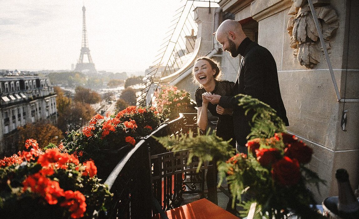 Пара смеется на балконе гостиницы Hotel Plaza Athenee на фоне Эйфелевой башни.