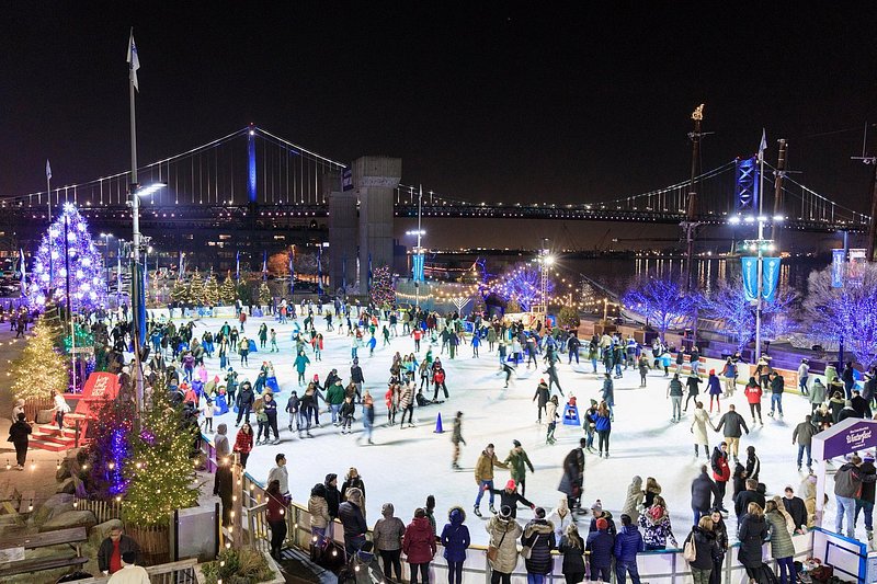 A crowd skates at night at the BlueCross River Rink in Philadelphia, with the Benjamin Franklin Bridge lit up in the background