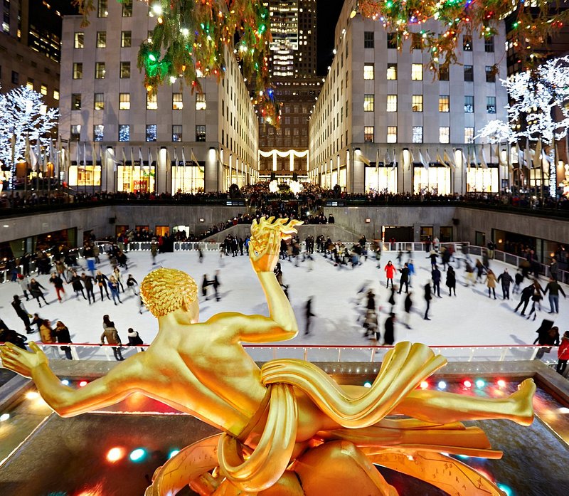 A golden statue is in the foreground, with ice skaters in motion at the Rockefeller Center skating rink