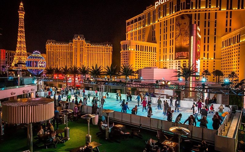 People are skating on an illuminated light blue swimming pool, with orange Las Vegas buildings, including a replica Eiffel Tower, lit up in the background 