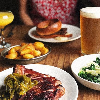 Disposição de pratos numa mesa no Marksman com uma mulher sentada em segundo plano. Os pratos incluem um prato de carne, verduras, batatas e um aro de cebola.