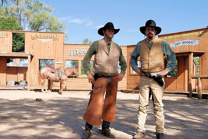 Dois cowboys com as mãos nas ancas com a fachada de um cenário ao estilo do Velho Oeste atrás