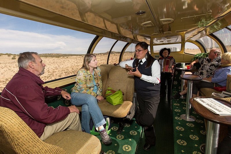 A steward having a conversation with passengers with tan, desert dirt visible through the floor to ceiling windows