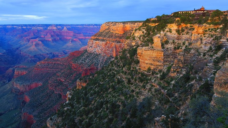 A Grand Canyon cliff at sunset, with a lit-up building at the top of the cliff. The cliffs are tan, deep red, and pink, and there are dark green shrubs dotting them.