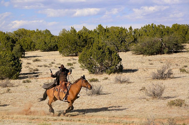 A cowboy robber rides a horse alongside the train among shrub trees and pale grasses.