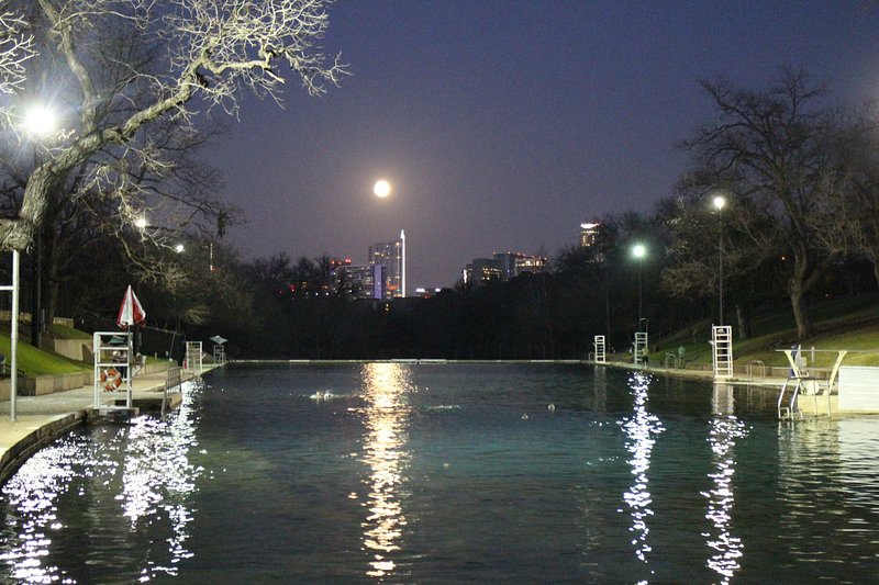 A full moon rises over the city of Austin, with the dark sparkling pools of Barton Springs in the front