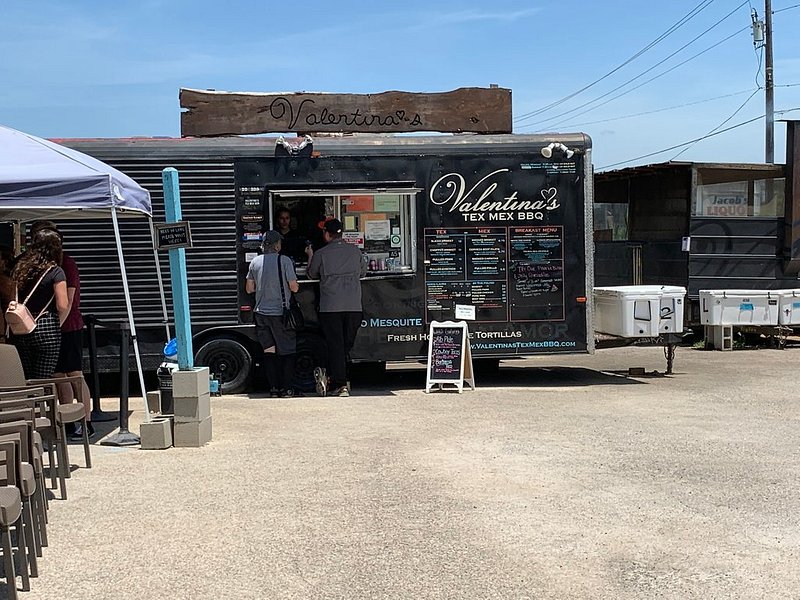 Two customers stand outside a black Valentina's Tex Mex BBQ food truck, with seating and a shade canopy to their left