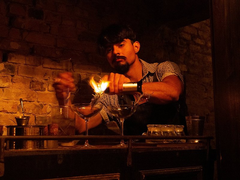 A bartender in a dark bar lights a cocktail with a piece of rosemary on the rim