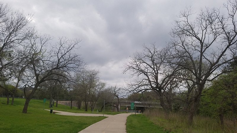 A sidewalk winds among grass and bare trees with a cement bridge in the distance an an overcast sky