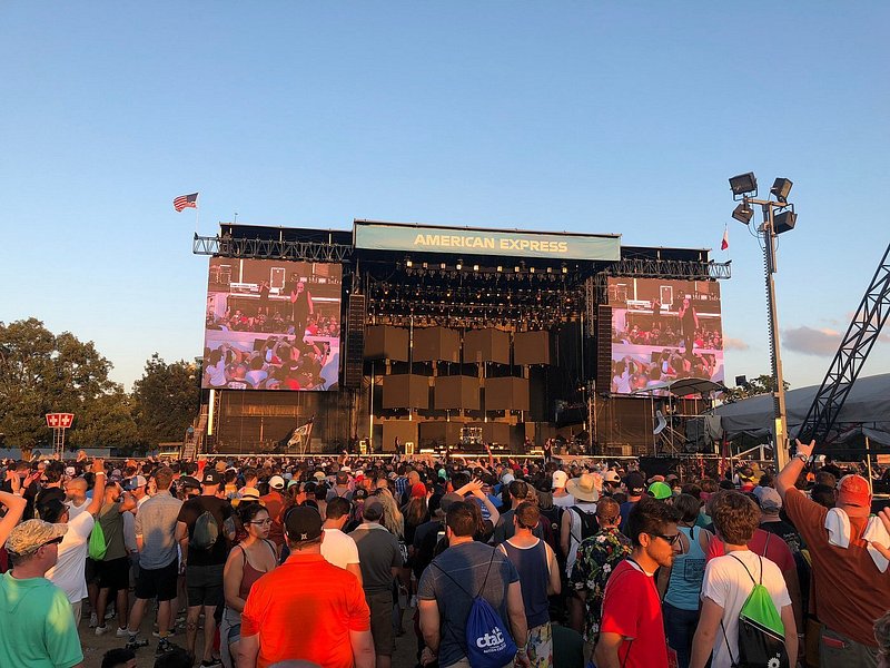 A crowd surrounds a stage with a large American Express sign hanging from the top, the light is soft pink on the stage suggesting sunset, but the sky is blue