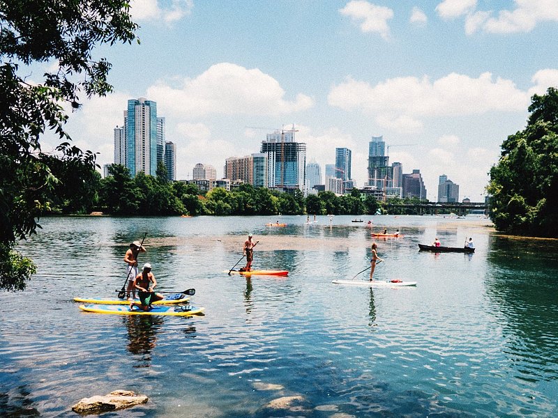 Paddle boarders glide across a reflective blue lake with the Austin skyline in the background with fluffy clouds dotting a blue sky