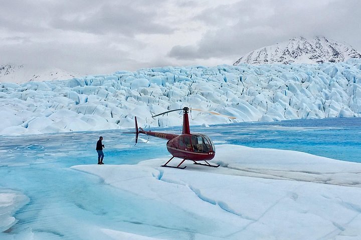 A person walks toward a red helicopter which is resting on a large glacier, with water channels carved into the ice behind it