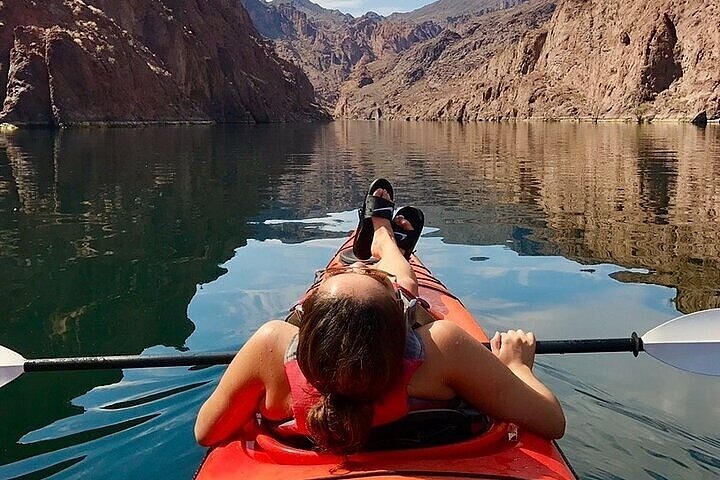 A woman reclines on an orange kayak in still, reflective waters, surrounded by canyon walls