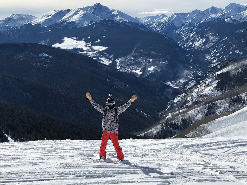 A skier stands facing the camera with their arms raised, a layer of packed snow beneath them and grey-blue mountains capped with snow in the distance behind them