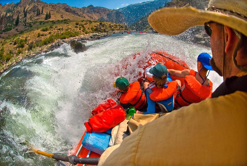 A close-up view of several rafters, one with a tan cowboy hat and three with bright orange life vests, as they enter gushing white water