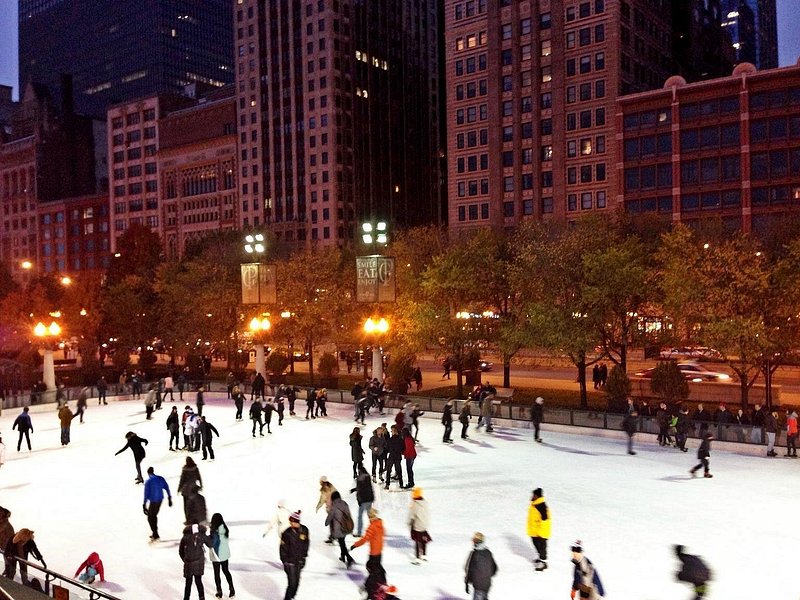 An evening crowd of ice skaters on a patch of white ice with dimly lit Chicago high rises in the background