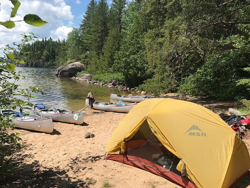 A yellow MSR tent sits on a sandy bank front of four canoes pulled just off of the shoreline, with large evergreen trees and rocks in the background