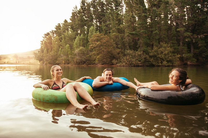 Three young people on inner tubes float on a calm lake with large trees in the background