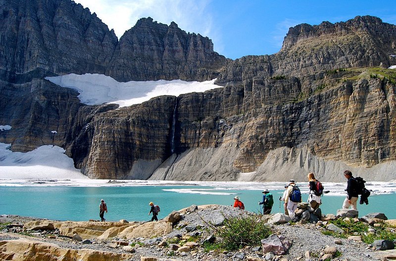 Eight hikers traverse rocky lands in front of a turquoise-blue glacial lake, with glaciers looming from cliffs in the distance