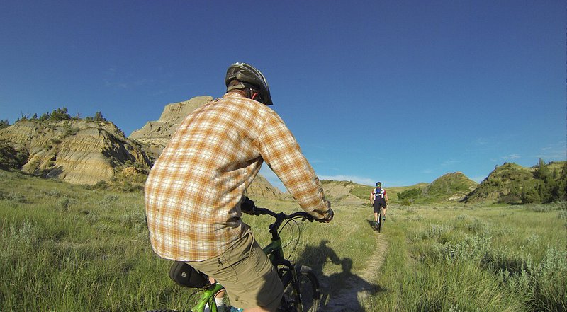 Up close to a mountain biker wearing a checkered shirt and helmet, with another one in the distance surrounded by light green grass and small hills
