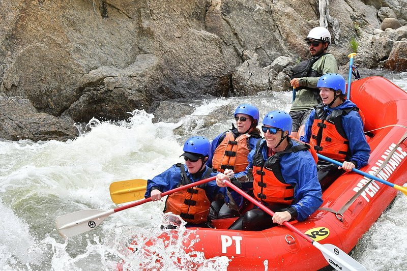 Five people in bright orange life jackets and blue helmets smile as they take on a whitewater section of river in an orange raft