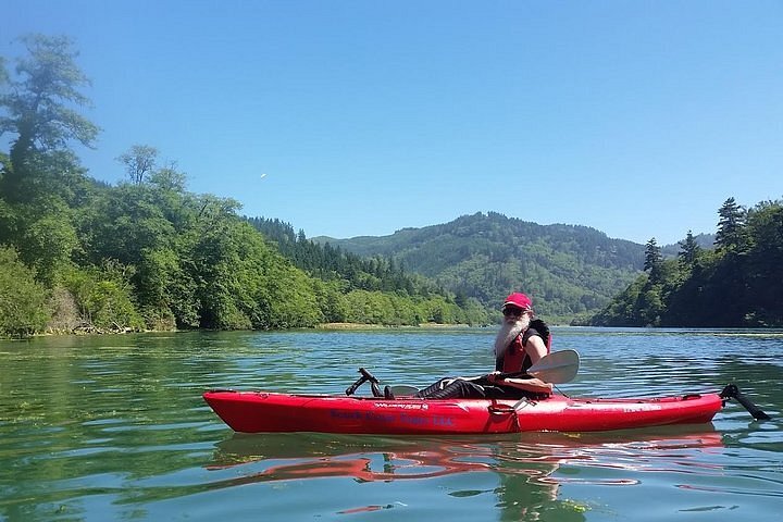 A bearded person in a red kayak on calm water looks at the camera with tree-lined hills in the background