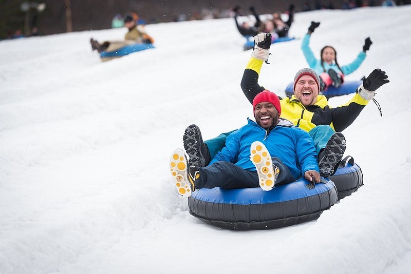 Two smiling adults slide down a snowy mountain side on a snow tube