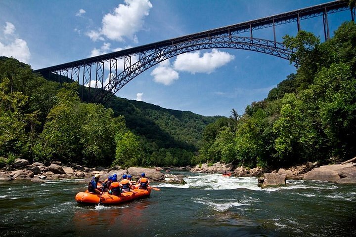 Rafters in a bright orange raft approach a small bit of white water and rocky shores. There's a large bridge looming hundreds of feet above them.