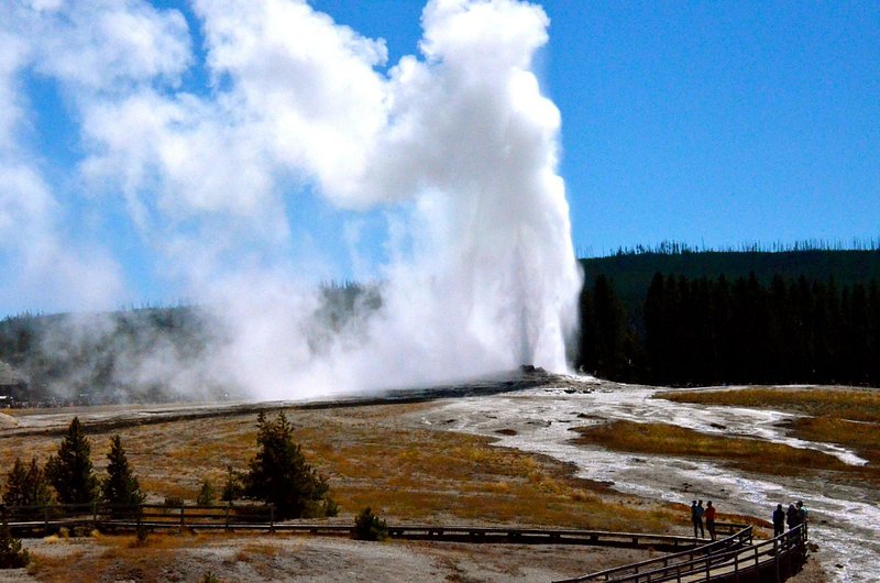 The Old Faithful geyser gushes into the air, clouds of mist wafting toward the left of the frame, and people stand on a board walk watching