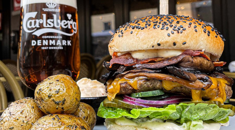 Gourmetburger med friskhakket oksekød, bacon, dobbelt ost, hjertesalat, svampe, agurk, rødløg, pickles, cremet dressing og hvidløgssmør. Serveres med ovnstegte kartofler og chilimayonnaise.