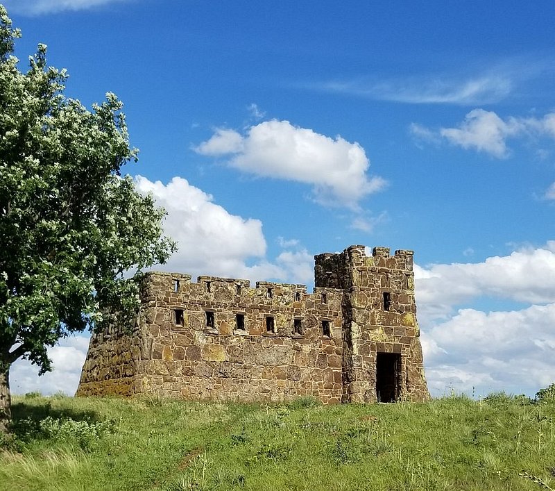 A small tan-colored stone castle sits on a grassy hill next to a large tree on a blue sky day with a few fluffy white clouds in the background