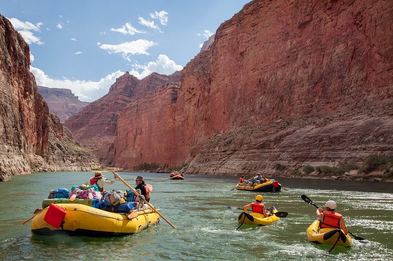 People move down river in a large yellow raft on the left, and two yellow kayaks on the right, with more rafts up a head. Around them are the rust-colored walls of the Grand Canyon 