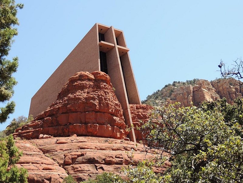 A cross leans out of a rectangular building among rust-colored rocks and desert-green foliage