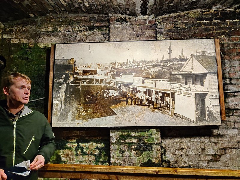 A blonde tour guide explains the history of the Seattle underground in front of a black and white photo on a brick wall