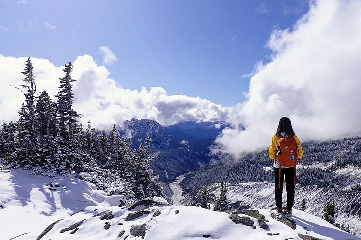 A person with long, dark hair and a bright yellow jacket and red backpack stand in front of a snow-covered landscape