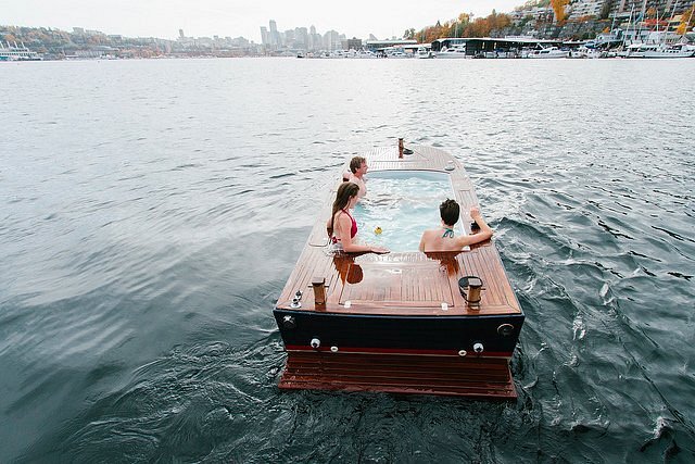 Three people sit inside a hot tub boat with the Seattle skyline in the far distance